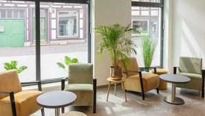 Een modern zithoekje met comfortabele stoelen en ronde tafels, grote ramen met uitzicht op de straat, en potplanten voor decoratie.