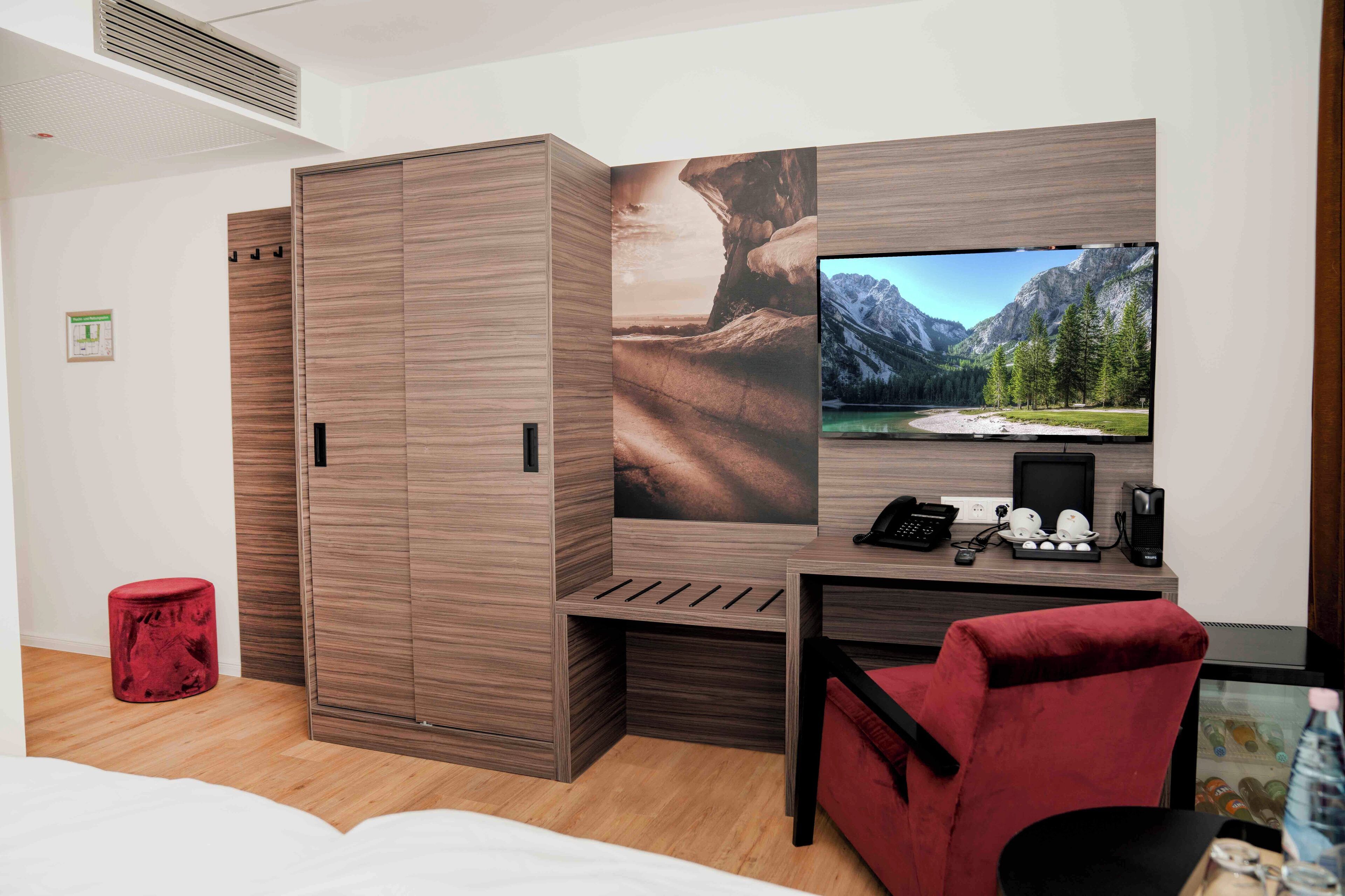 Une chambre d’hôtel avec un lit, un bureau avec un ordinateur, une chaise rouge, une armoire et un grand téléviseur diffusant un paysage de montagne.