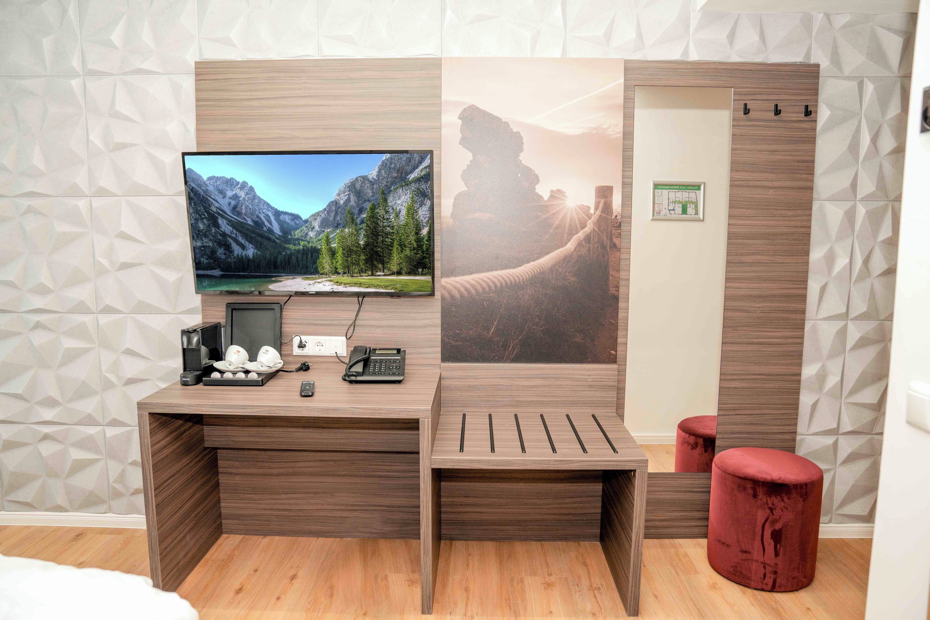 Quarto de hotel moderno com escrivaninha de madeira, televisão de tela plana, um banquinho vermelho e um pôster grande de uma paisagem montanhosa.