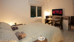 Una habitació d'hotel amb un llit, escriptori, cadira, televisió que mostra 'NETFLIX' i finestra amb persianes.