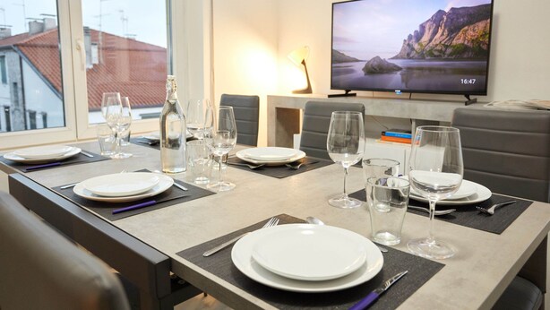 Zona de dinar amb taula preparada per a quatre, televisió que mostra un paisatge idíl·lic i vista cap a edificis a través de la finestra.