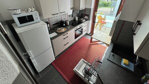 Cocina compacta con gabinetes blancos, microondas y cafetera. Un tapete rojo cubre el suelo, y una ventana da a un espacio exterior con una silla.