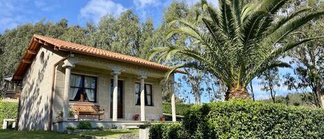 A house with a tiled roof, a front porch with a bench, and a well-maintained garden with a palm tree.