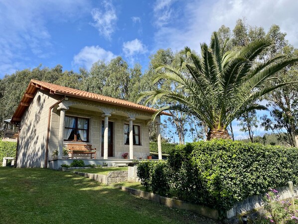 A house with a tiled roof, a front porch with a bench, and a well-maintained garden with a palm tree.