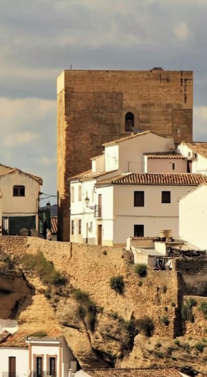 A historic stone tower with a flat roof, adjacent white buildings with tiled roofs, and a hillside with sparse vegetation.