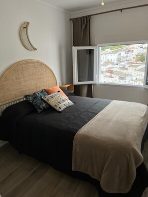 A bed with a curved headboard, a window with a view of buildings, and a bedside table with a lamp.