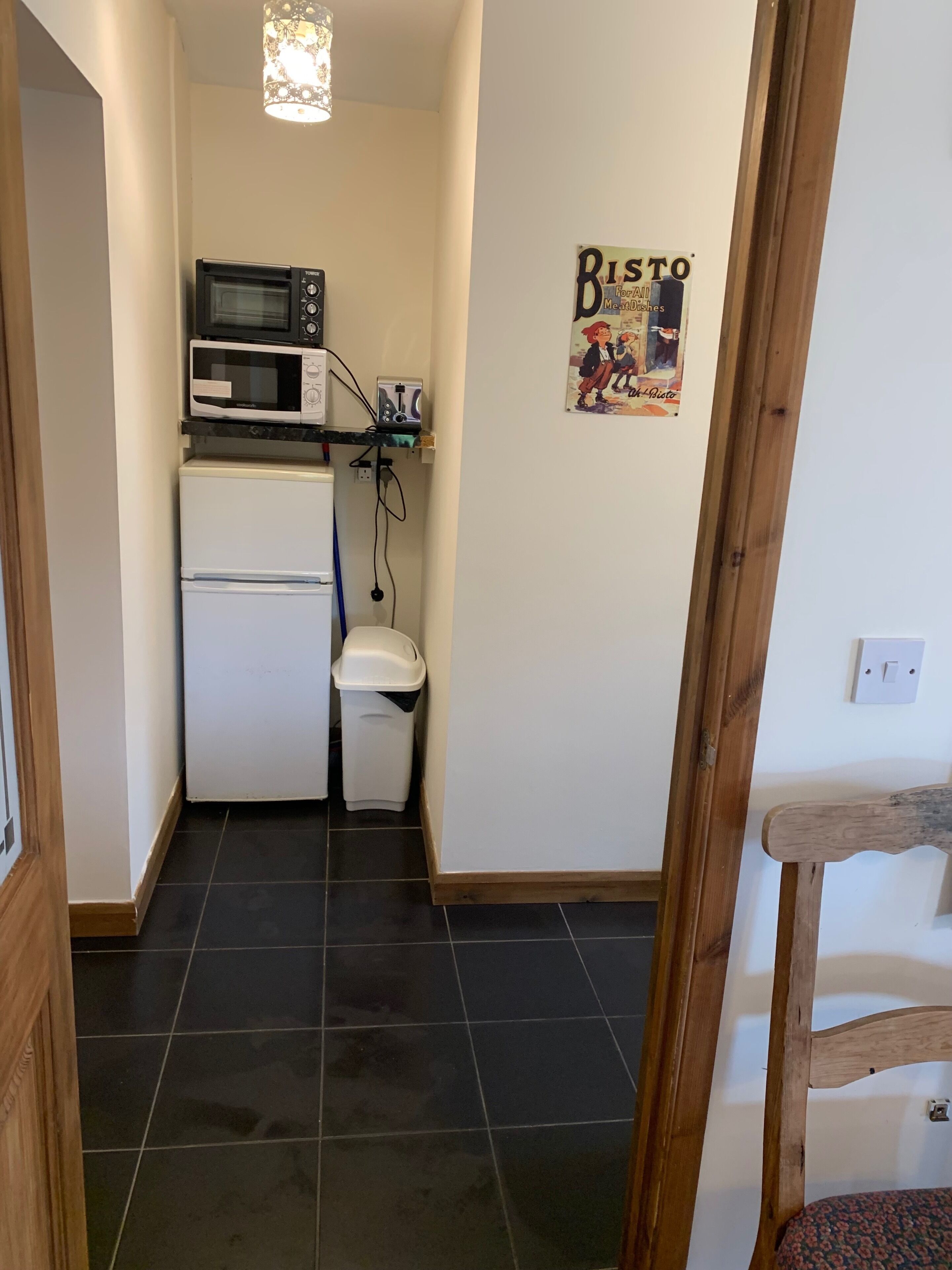 Una cucina compatta con forno a microonde, tostapane e frigorifero. Si nota un cestino e una locandina appesa al muro.