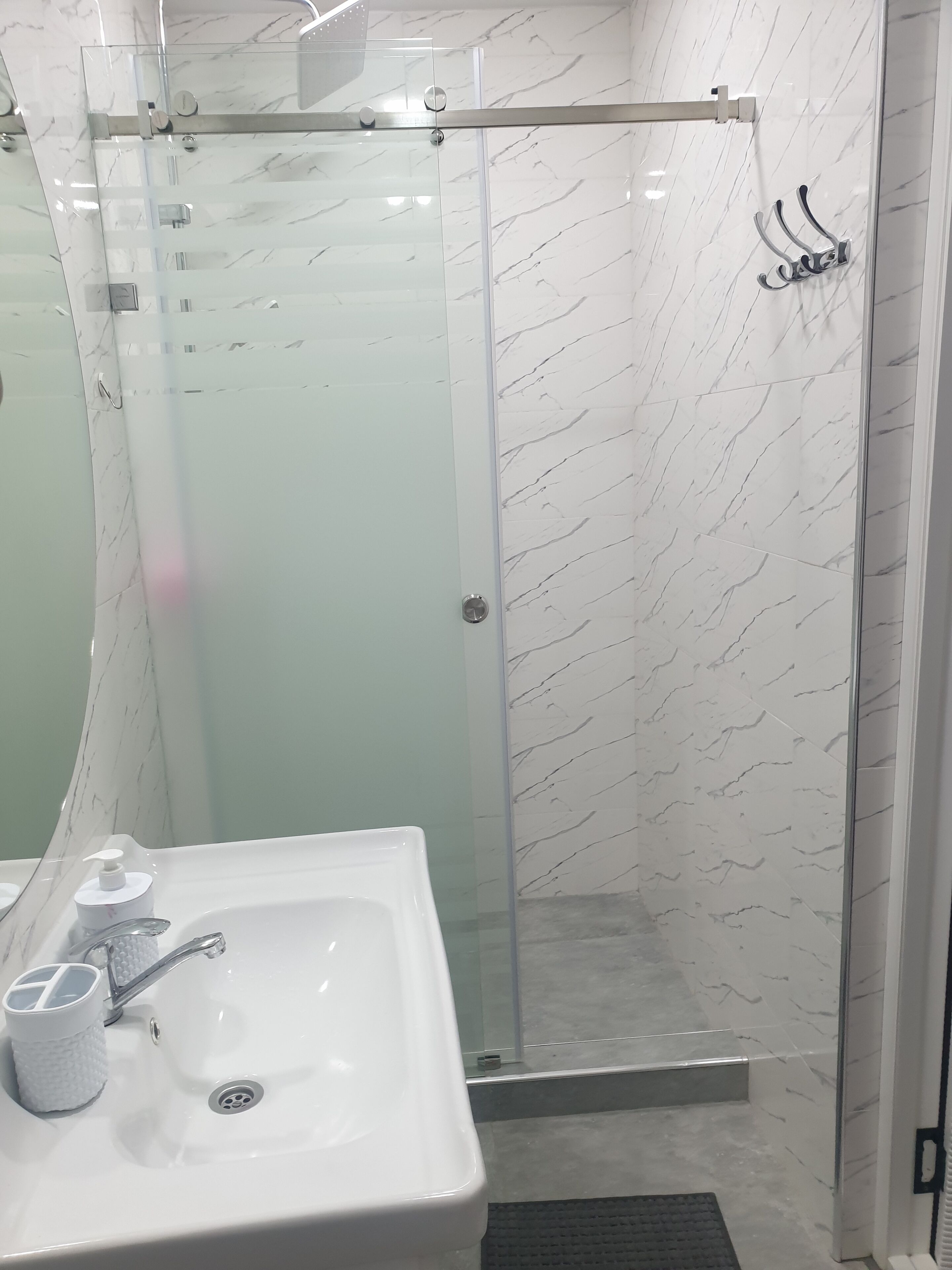 Un baño moderno con ducha de vidrio, lavamanos blanco con dispensador de jabón y pared con estampado de mármol.