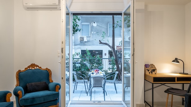 Een moderne woonkamer met een blauwe fauteuil, een houten bureau en een glazen deur die uitkomt op een terras met een tafel en stoelen.