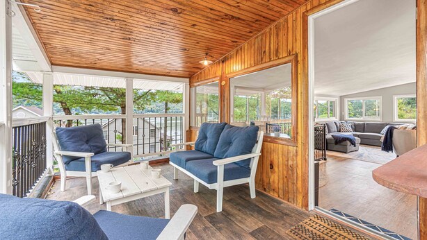 Un espace extérieur abrité, doté d’un plancher en bois, de mobilier blanc et de grandes fenêtres offrant une vue sur la verdure environnante.
