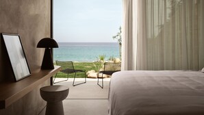 Een slaapkamer met een groot raam dat uitkijkt op de oceaan, een bed, een stoel, een tafel en een lamp.