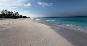 A wide sandy beach with gentle waves, clear skies, and sparse vegetation.
