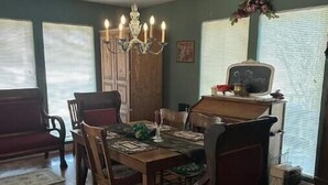 A dining room with a wooden table set for four, surrounded by chairs. A chandelier hangs from the ceiling, and there is a cabinet with a mirror above the table.