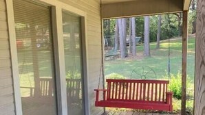 A wooden porch with a red swing, a sliding glass door, and a view of a grassy yard.
