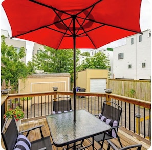 Um terraço com mesa e cadeiras de vidro, um guarda-sol vermelho e vista para as casas vizinhas.