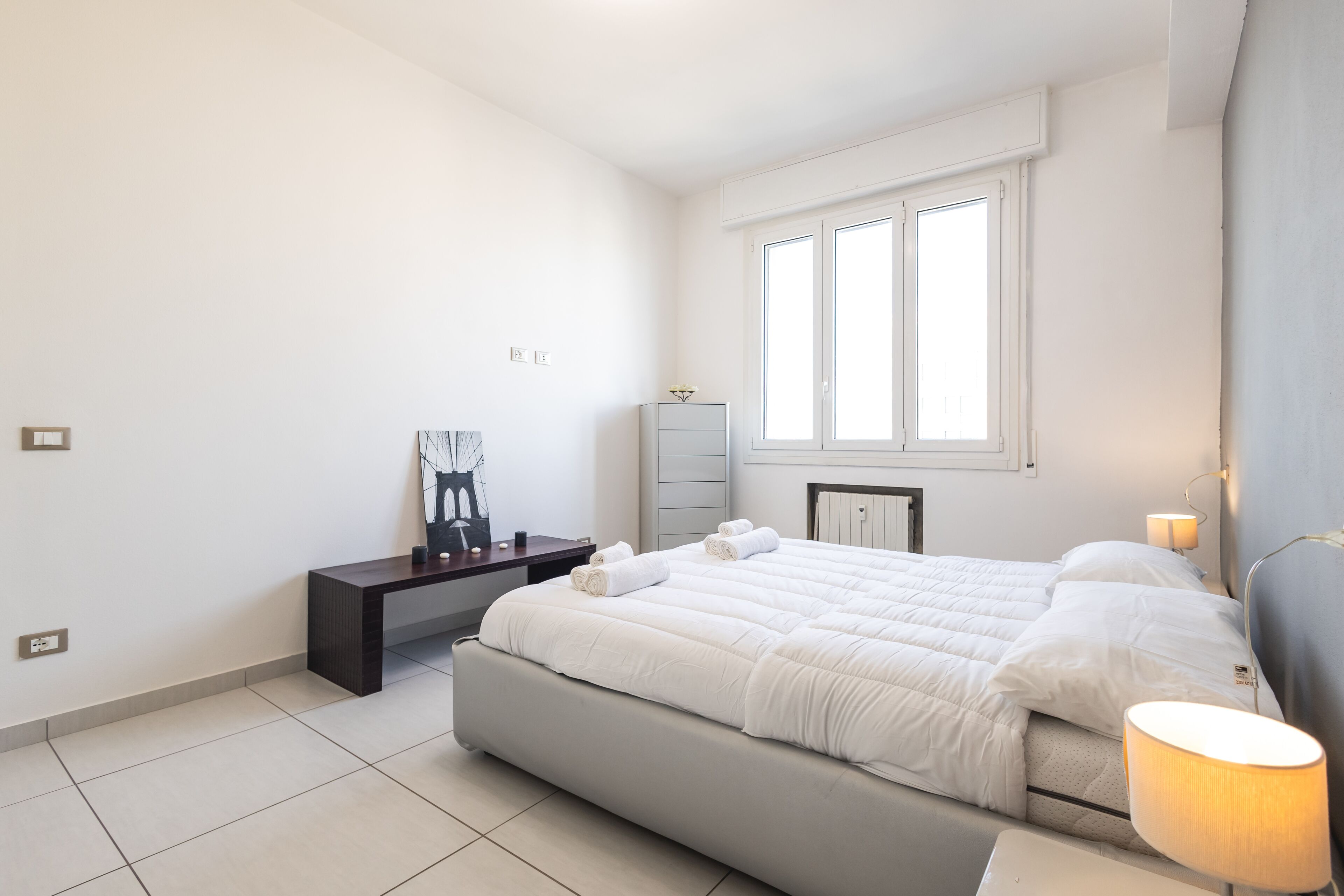 A modern bedroom with a large bed, a bedside table, a desk, and a window.