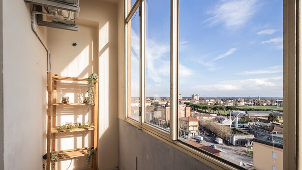 A room with a large window offering a city view, a wooden shelf with plants, and a visible air conditioning unit. - Ravenna Sky View Apartment (Ravenna)