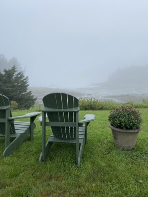 Two green Adirondack chairs and a potted plant with red flowers on a grassy area.