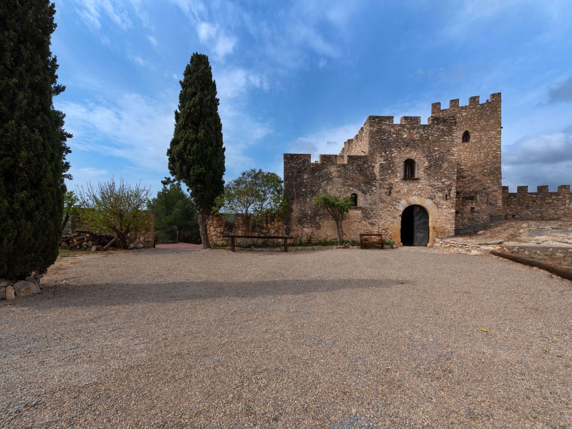 Un bâtiment historique en pierre, doté d’une grande entrée en arc de cercle, entouré d’une cour recouverte de gravier et d’un grand arbre.