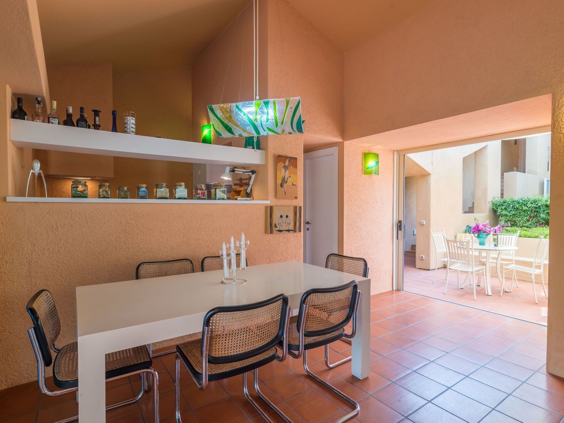 A dining area with a white table and chairs, a hanging light fixture, and a shelf with bottles.