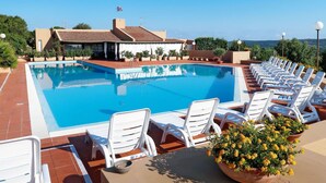 A swimming pool with white lounge chairs and a building with a flag in the background. - Cosy apartment in villa for 5 guests with A/C, pool, WIFI, TV, terrace and panoramic view (Palau)