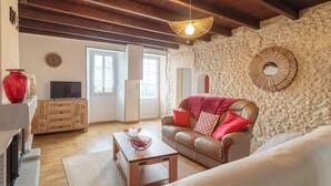 Un salon chaleureux avec un mur en pierre, un plafond en bois, un canapé en cuir agrémenté de coussins rouges, une table basse en bois et un téléviseur posé sur une console en bois.