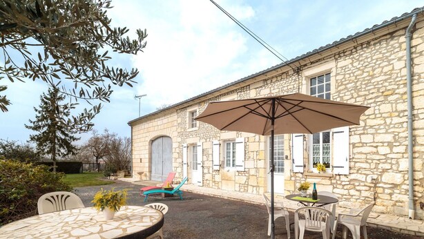 Une terrasse extérieure avec une table et des chaises en pierre, un grand parasol et un bâtiment en pierre doté de fenêtres et de volets.