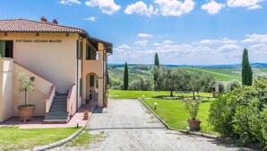 Un bâtiment appelé « Fattoria Luciano Mazzei », doté d’un escalier menant à l’entrée, entouré d’un jardin bien entretenu et offrant une vue dégagée sur la campagne.