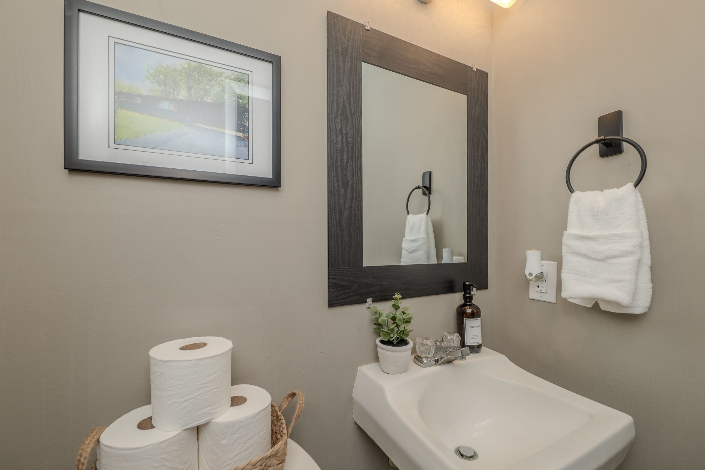 Une salle de bain avec un tableau encadré, un grand miroir, un lavabo et deux rouleaux de papier toilette.