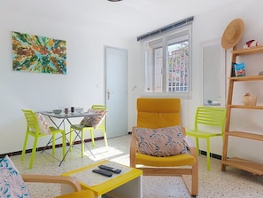 Een kamer met een eettafel, stoelen, een gele fauteuil, een boekenkast, een schilderij en een raam met uitzicht.
