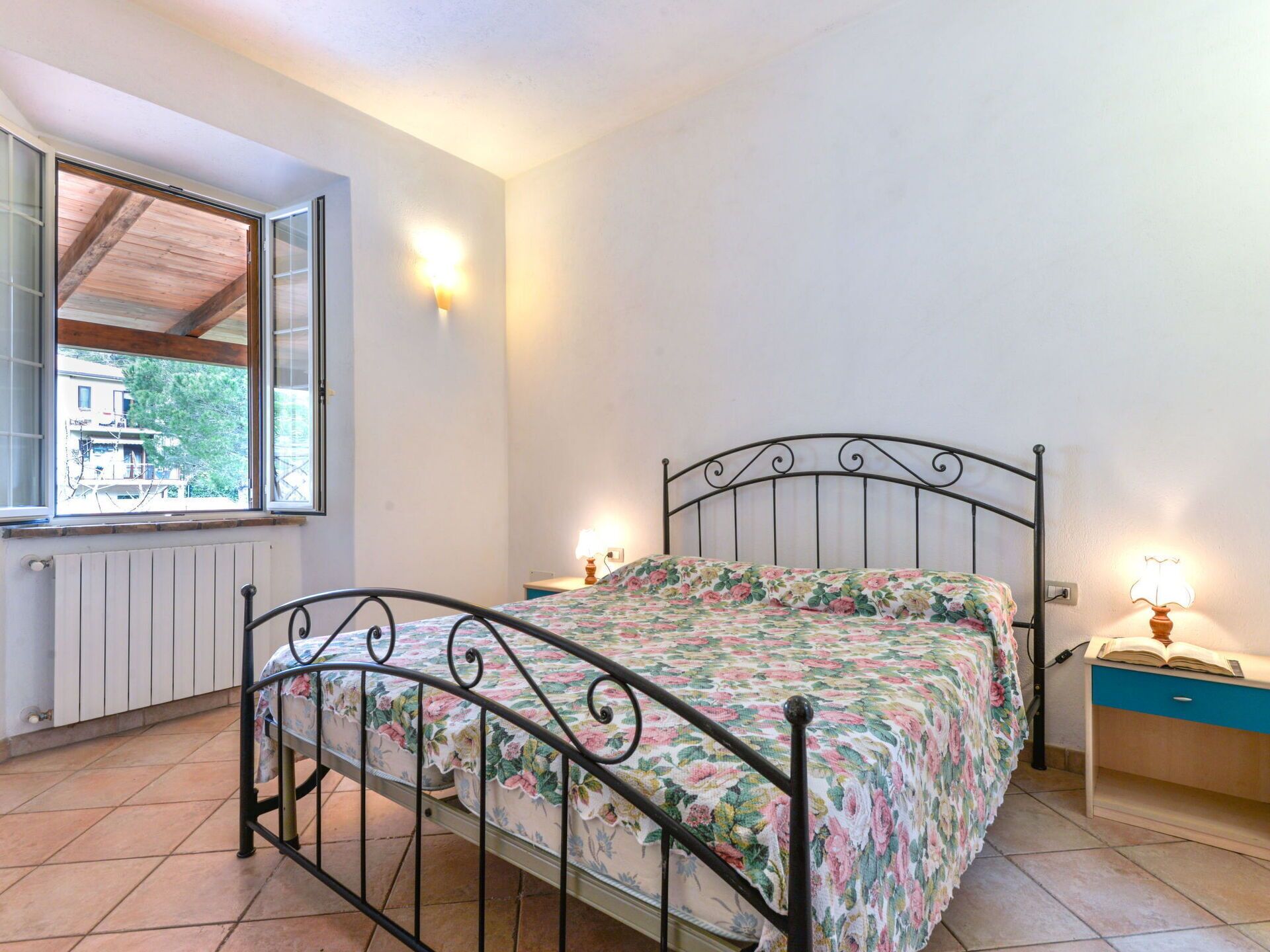 Un dormitorio con estructura de cama metálica, ropa de cama floral, una ventana con vistas y lámparas de noche.