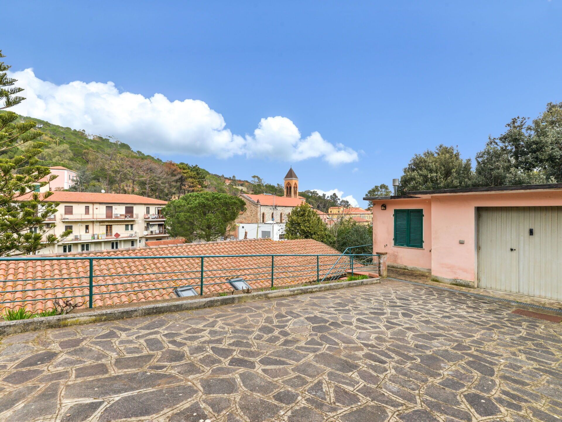 Un patio empedrado con techo de tejas, un pequeño edificio con un persiano verde y una iglesia con campanario al fondo.