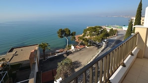 Une vue sur la côte, depuis un balcon donnant sur une rue bordée d’arbres et sur une mer bleue et limpide.