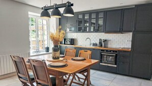 A modern kitchen with dark cabinets, a wooden dining table, and pendant lights. - Beautiful  villa for 3 people with WIFI, TV and patio (Petit Saint-malo)