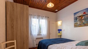 A bedroom with a wooden ceiling, a bed with white and blue bedding, a chair, a window with sheer curtains, and a painting on the wall.
