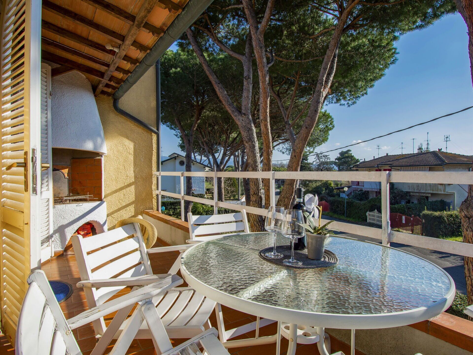 A covered outdoor seating area with a round glass table and white chairs, a pizza oven, and a view of a residential area with trees and houses.