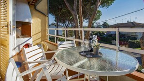 Un espacio de estar al aire libre techado con una mesa de vidrio redonda y sillas blancas, un horno de pizza, y vistas a una zona residencial con árboles y casas.