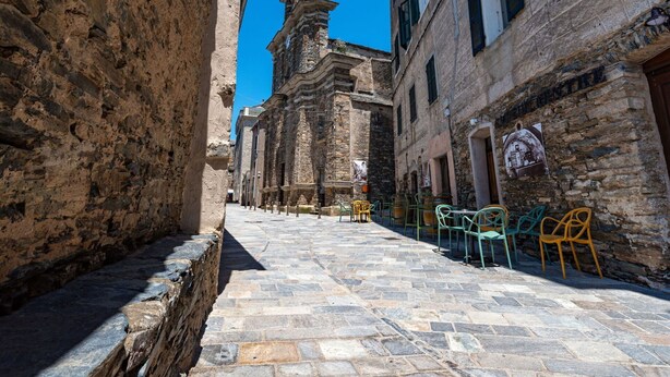 A narrow cobblestone street with stone buildings, a church tower, and outdoor seating with chairs.