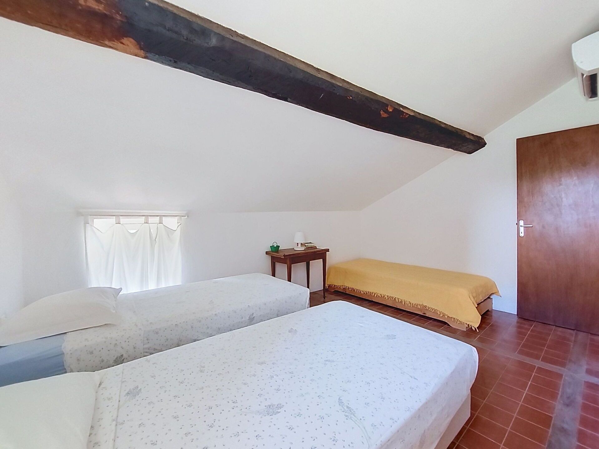 A room with two beds, a window with curtains, a wooden door, and a small table with a green basket.