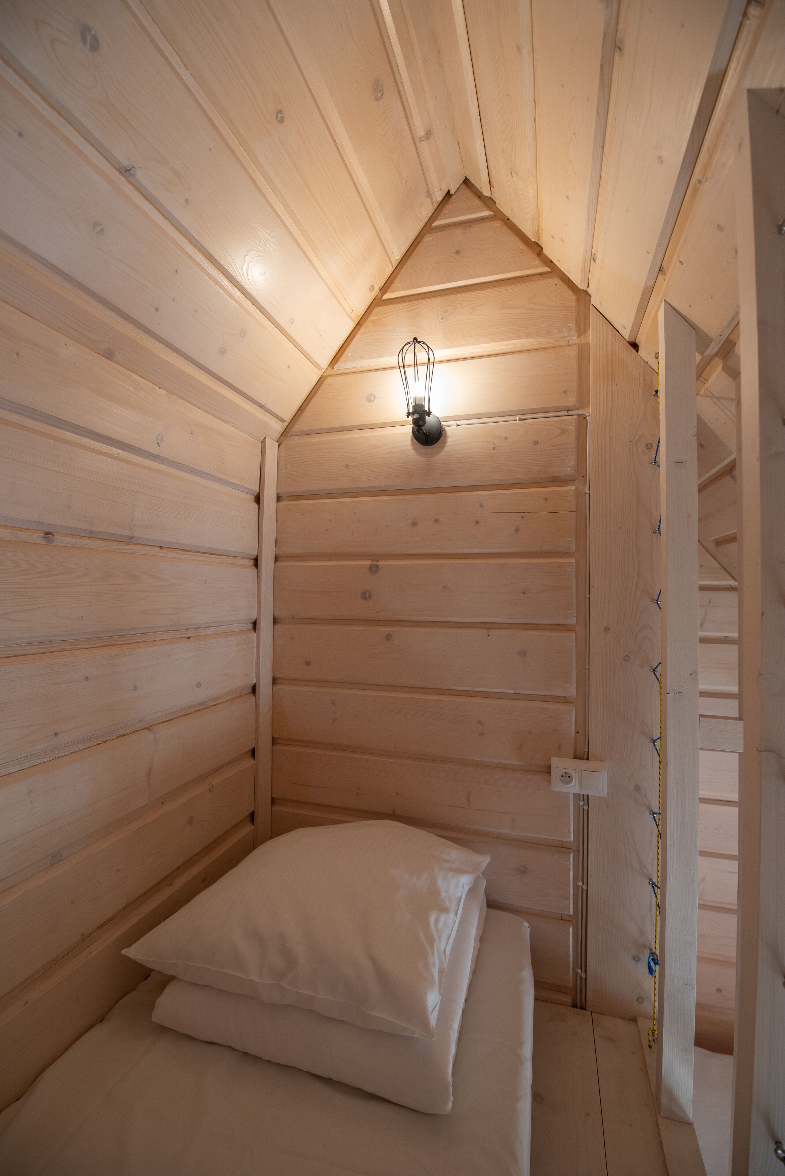 Ein kleines, gemütliches Holzhütten-Zimmer mit schrägverdeckter Decke, einem Einzelbett mit weißer Bettwäsche und einer wandmontierten Leuchte.