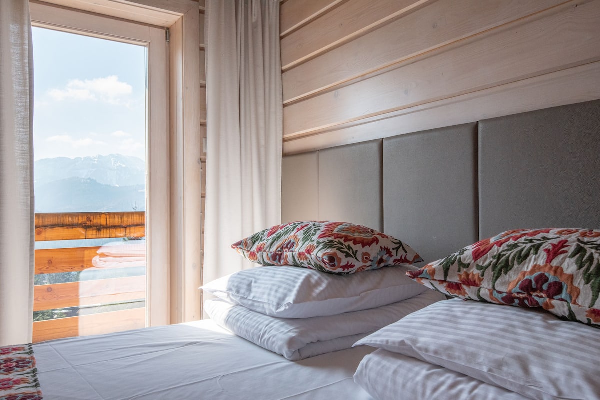 Ein Hotelzimmer mit Bett, Kissen, einem Fenster mit Vorhängen und Blick auf Berge.