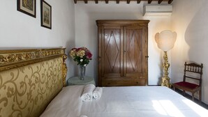 Une chambre à coucher avec un grand lit, une armoire en bois, une petite table sur laquelle est posé un vase de fleurs, une lampe et une chaise.