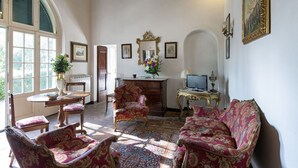 Une pièce spacieuse avec une grande fenêtre, un plafond en bois et une cheminée traditionnelle. On y trouve deux canapés richement décorés et une petite table sur laquelle est posé un vase de fleurs.