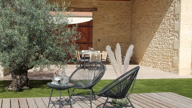 Een terras buiten met een houten terras, twee zwarte draadstoelen, een ronde tafel met een glazen blad, en een klein tafeltje met een vaas en kaars.