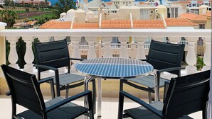 Een balkon met een ronde tafel en vier stoelen, met uitzicht op een kustplaatsje met gebouwen en een heldere lucht.