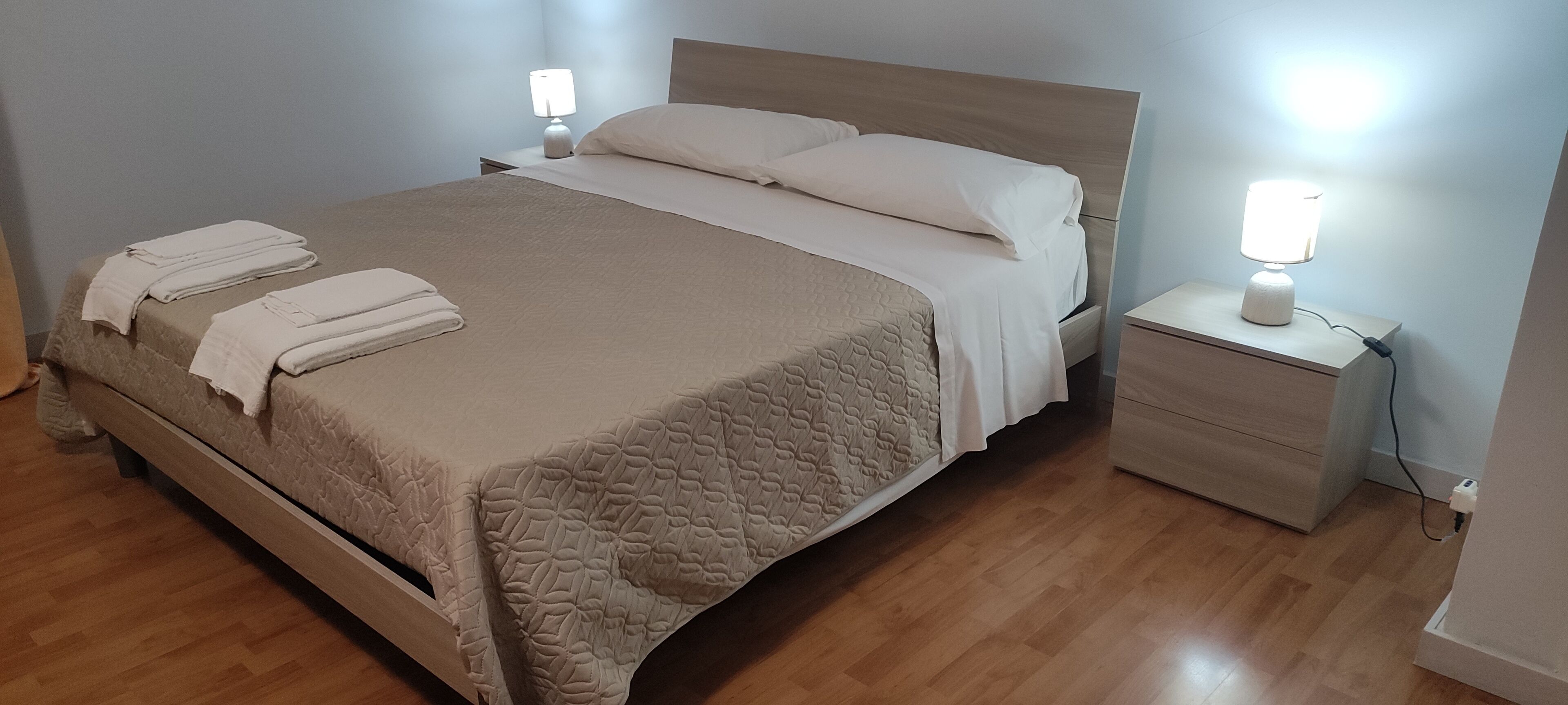 Un lit bien fait, recouvert d’une courtepointe beige, avec deux oreillers et des serviettes pliées, posé sur un plancher en bois. Il y a deux tables de chevet avec des lampes, une de chaque côté du lit.