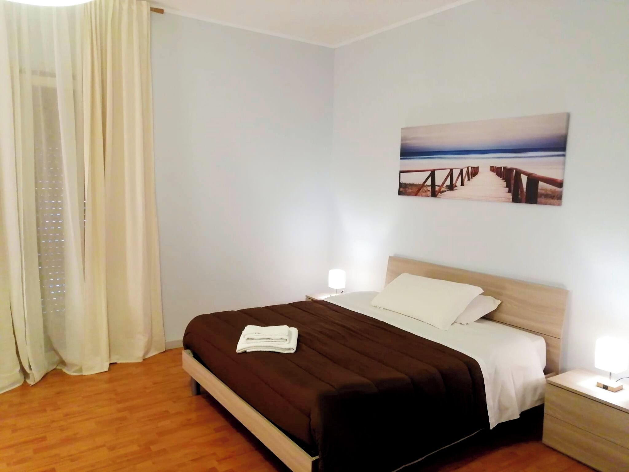 Siististi järjestetty hotellihuone, jossa on puulattia, ruskealla peitteellä ja valkoisilla tyynyillä varustettu sänky, yöpöytä lampun kanssa ja seinällä oleva taulu, jossa on kuvattuna laituri.