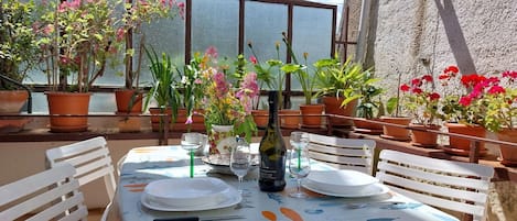Un área de comedor al aire libre con una mesa preparada para dos personas, rodeada de plantas en macetas y vistas al mar.