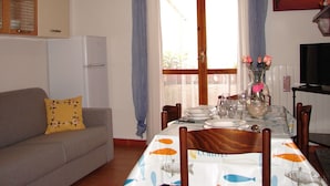 Ein Essbereich mit Tisch für vier Personen, eine Couch mit floralem Kissen, ein Kühlschrank und ein Elefantenbild.