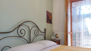 Ein Bett mit Metallkopfteil, weißen Kissen und einer gelben Tagesdecke. Ein kleiner Nachttisch mit Blumen in einer Vase und einer Uhr steht daneben. Ein Gemälde hängt an der Wand über dem Bett.
